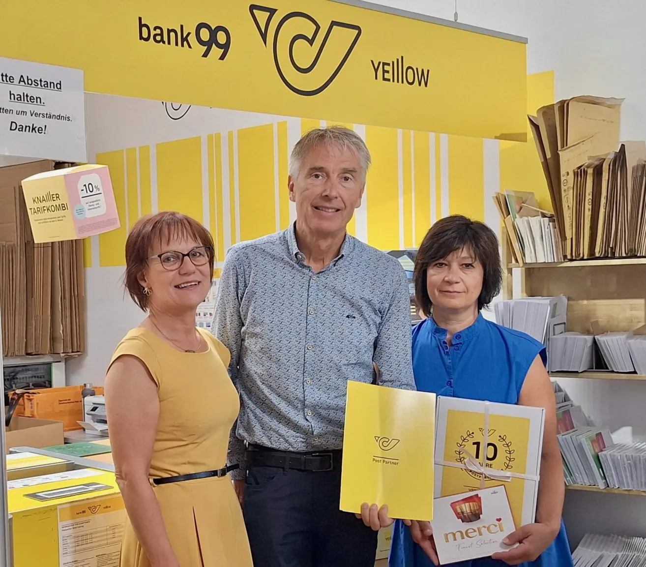 Drei Personen stehen vor einem gelben Banner mit den Texten 'bank99' und 'YELLOW'. Der Mann hält ein gelbes Buch mit dem Wort 'merci'. Die beiden Frauen tragen gelbe und blaue Kleider. Dahinter steht ein Regal mit Büchern.