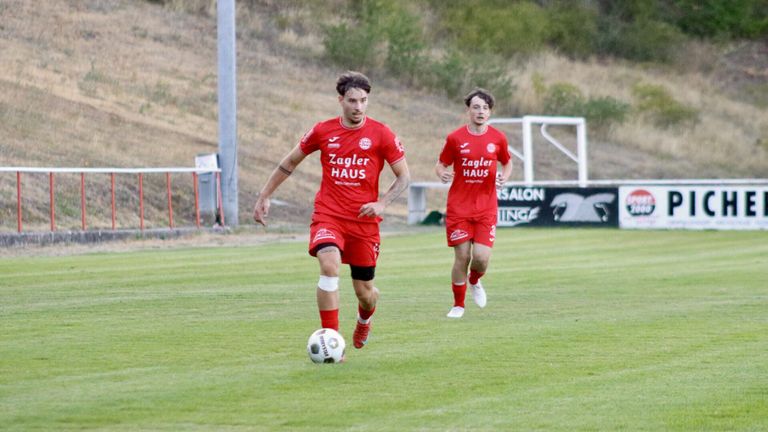 Zwei Fußballspieler in roten Trikots laufen auf einem Feld. Einer dribbelt einen Fußball, während der andere dicht hinter ihm herläuft. Das Feld hat eine Rasenfläche und ist von einem Zaun mit einem Banner umgeben.