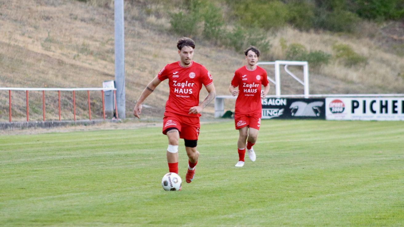 Zwei Fußballspieler in roten Trikots laufen auf einem Feld. Einer dribbelt einen Fußball, während der andere dicht hinter ihm herläuft. Das Feld hat eine Rasenfläche und ist von einem Zaun mit einem Banner umgeben.