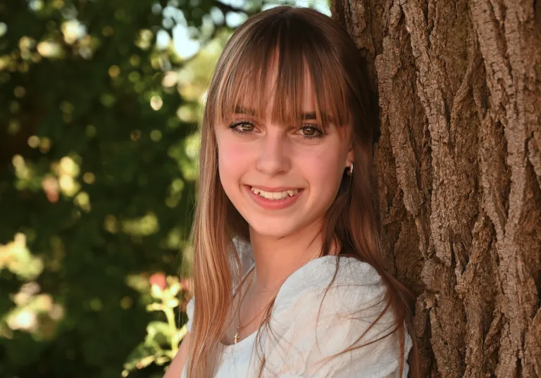 A smiling young woman with long brown hair and bangs is standing next to a tree trunk.