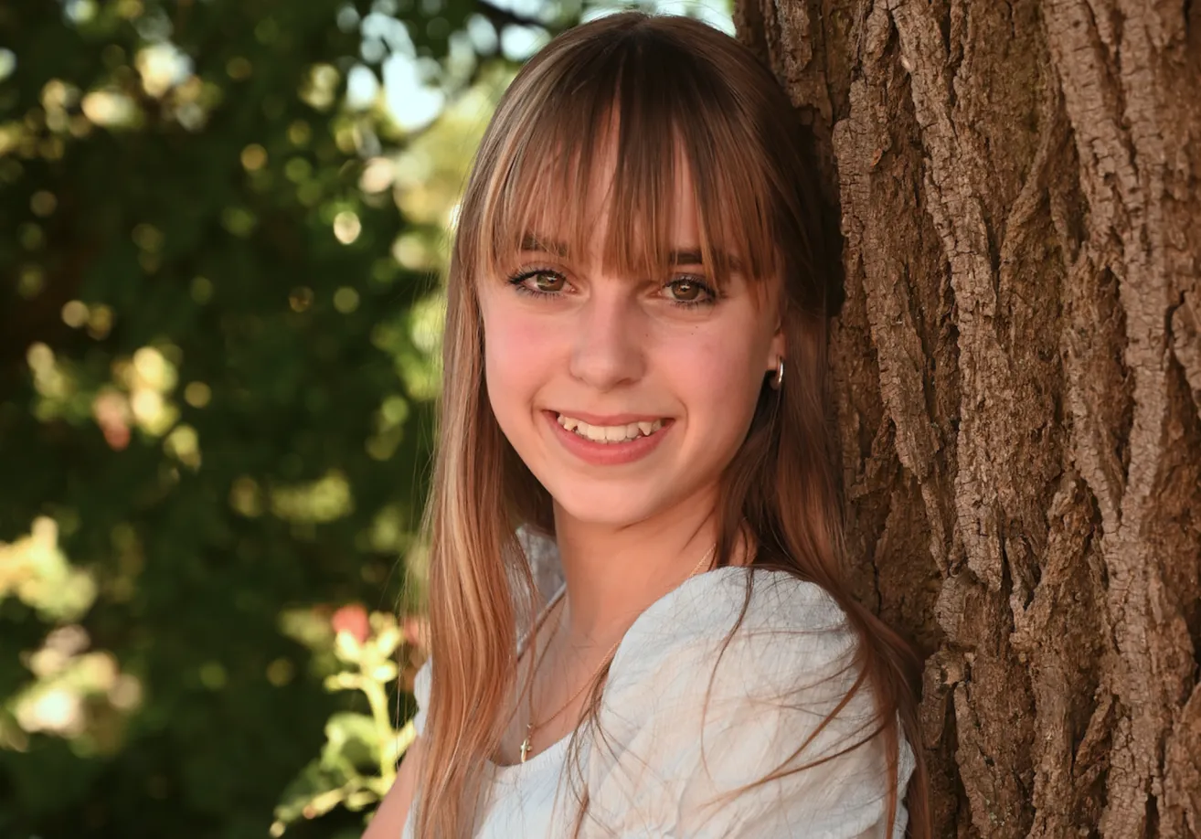 A smiling young woman with long brown hair and bangs is standing next to a tree trunk.