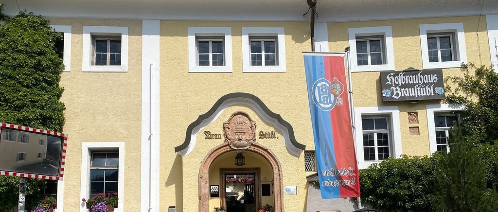 Die Vorderseite eines gelben Gebäudes mit weißen Verzierungen, mit einem großen gewölbten Eingang, einer Flagge und mehreren Fenstern. Der Eingang hat eine dekorative Plakette darüber.