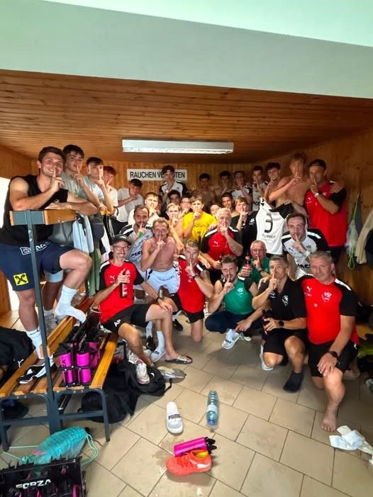 Eine Gruppe von Fußballspielern in einer Umkleidekabine, einige sitzen auf einer Bank, andere stehen, alle tragen Trikots und Shorts und posieren für ein Foto. Der Raum hat Holzwände und eine Deckenleuchte.