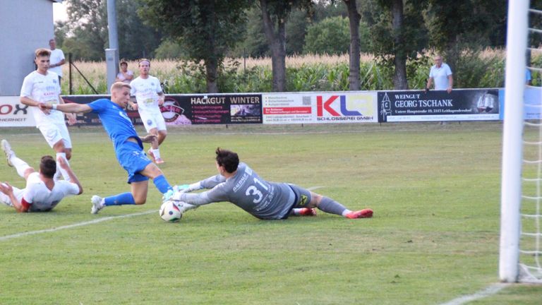 Fußballspieler auf einem Feld, ein Spieler in Blau versucht, den Ball zu schießen, während ein anderer in Grau auf dem Boden liegt und versucht, den Schuss zu blockieren.