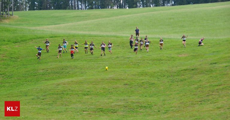 A group of people are running on a grassy field, possibly in a race. They are wearing numbered jerseys and sneakers. A yellow object is on the ground. In the distance, there are trees.