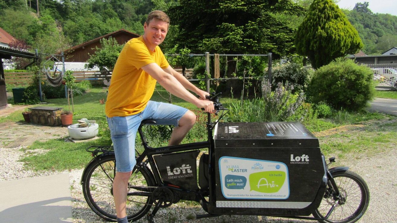 Ein Mann in einem gelben T-Shirt und Jeansshorts fährt ein Lastenrad mit einem schwarzen Kasten vorne, der mit 'Loft Kreativagentur' beschriftet ist. Er lächelt und posiert für ein Foto im Garten.