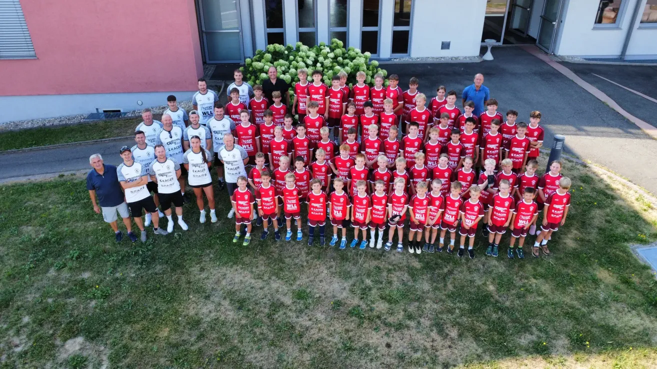 A group photo of a sports team, including children and adults, wearing red and white jerseys, standing in a line on a grassy field.