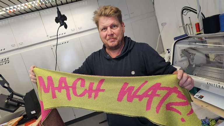 Ein Mann in einem schwarzen Hoodie hält ein gestricktes Banner mit rosa Buchstaben, auf dem 'MACH WATTS' steht, vor einem weißen Schrank.