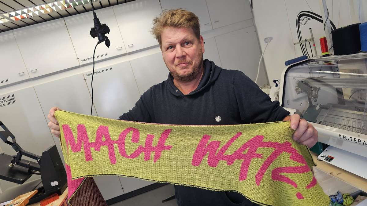 Ein Mann in einem schwarzen Hoodie hält ein gestricktes Banner mit rosa Buchstaben, auf dem 'MACH WATTS' steht, vor einem weißen Schrank.