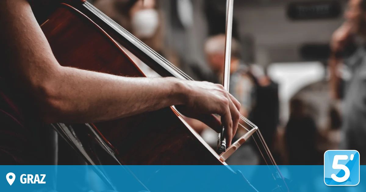 Ein Musiker spielt ein Cello und trägt eine Maske, mit einem verschwommenen Hintergrund anderer Musiker.