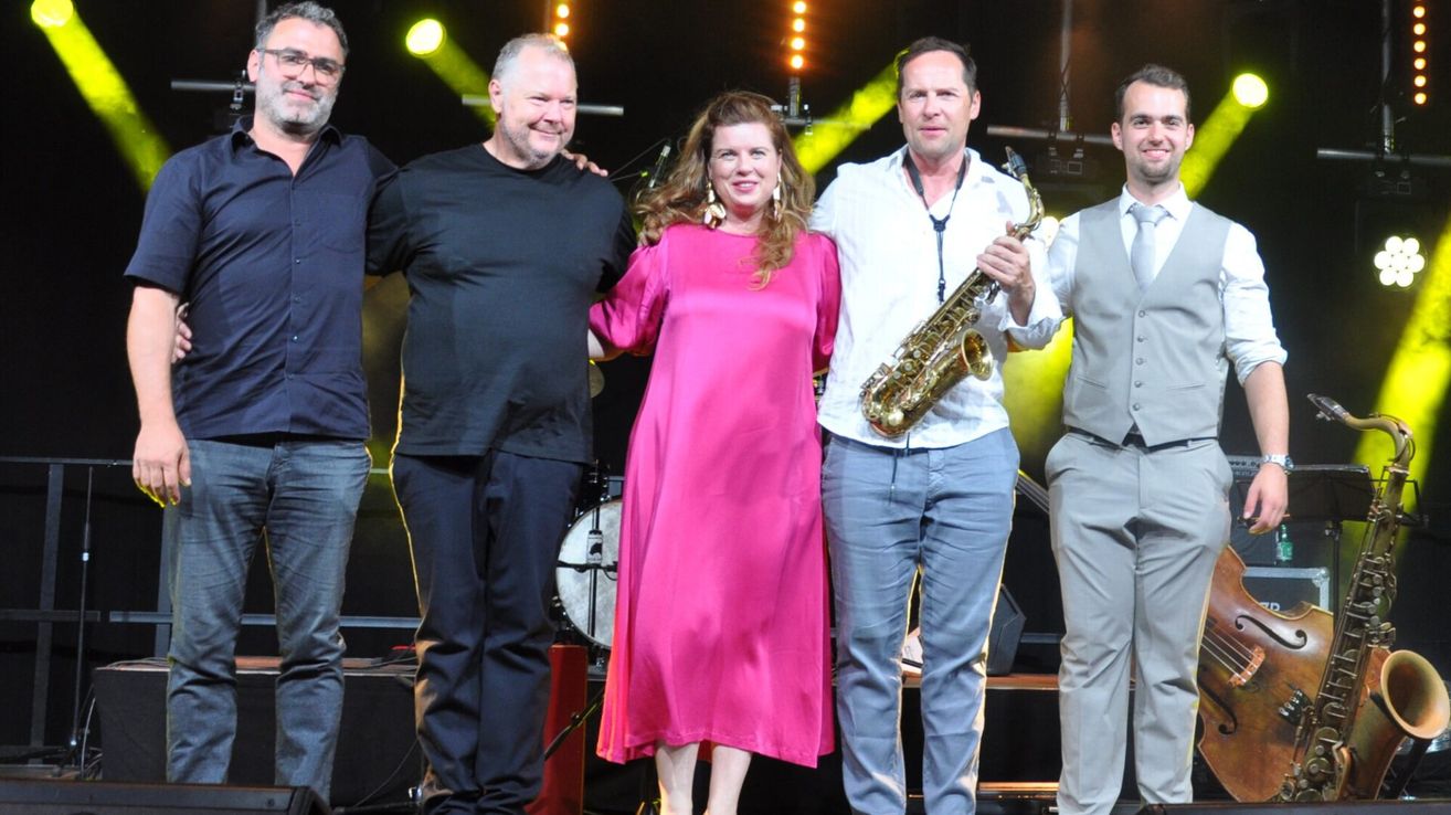 Eine Gruppe von Musikern auf der Bühne, lächelnd und für ein Foto posierend. Eine Frau in einem rosa Kleid steht zwischen zwei Männern, einer hält eine Saxophon. Hinter ihnen ist ein Schlagzeug-Set und Bühnenlichter.