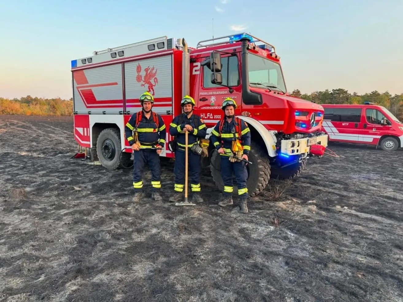 Drei Feuerwehrleute stehen vor einem roten Feuerwehrauto mit der Nummer 2 darauf und halten eine Schaufel und ein Werkzeug. Sie tragen Helme und Uniformen. Der Boden ist mit Asche bedeckt.