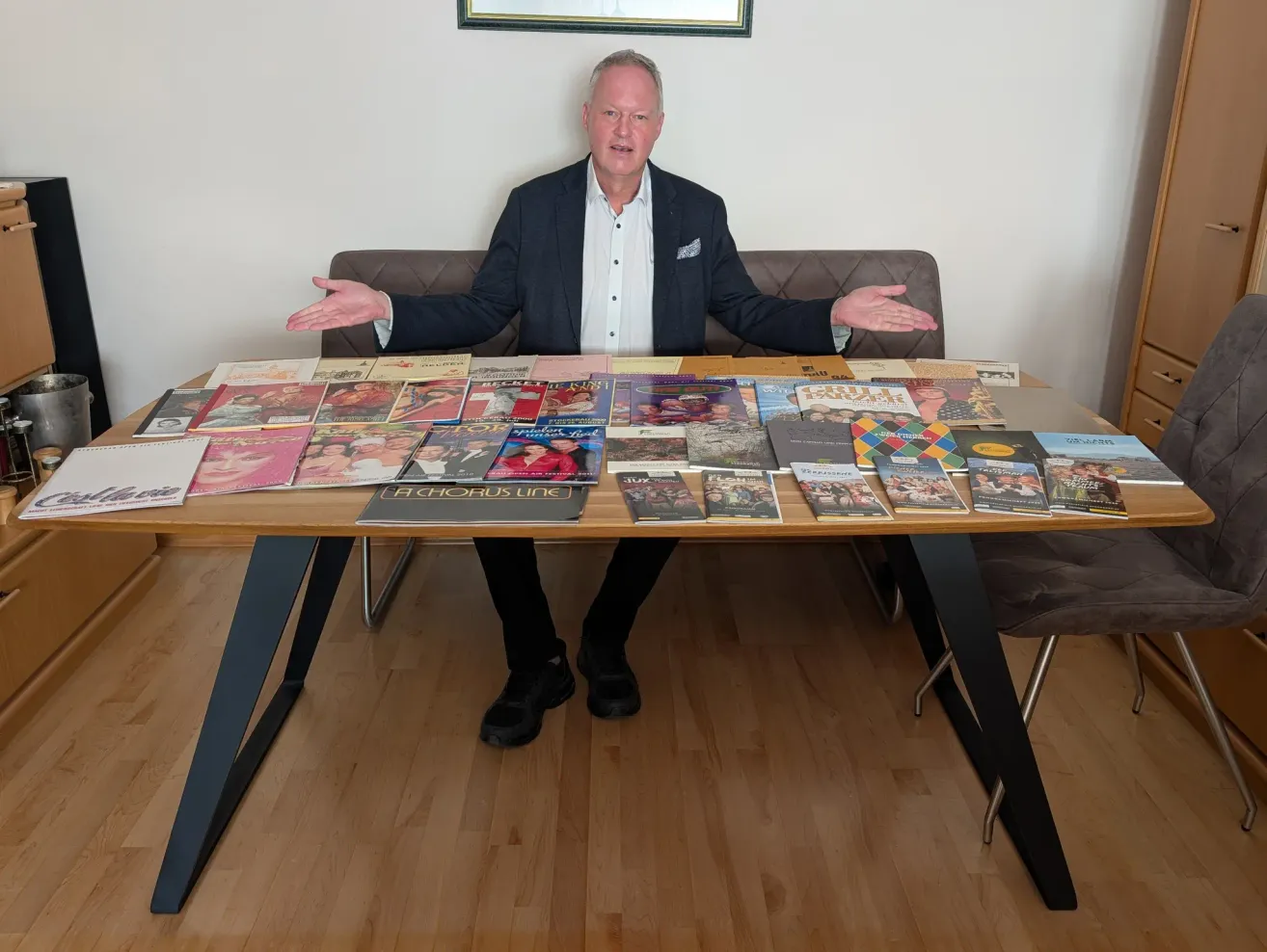 Ein Mann in einem Anzug sitzt an einem Tisch mit einer Sammlung von Zeitschriften und Büchern. Er gestikuliert mit den Händen und lächelt.