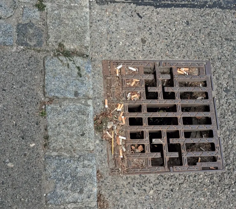 Eine Nahaufnahme eines schmutzigen Kanaldeckels mit Zigarettenkippen und Ablagerungen auf dem Boden.