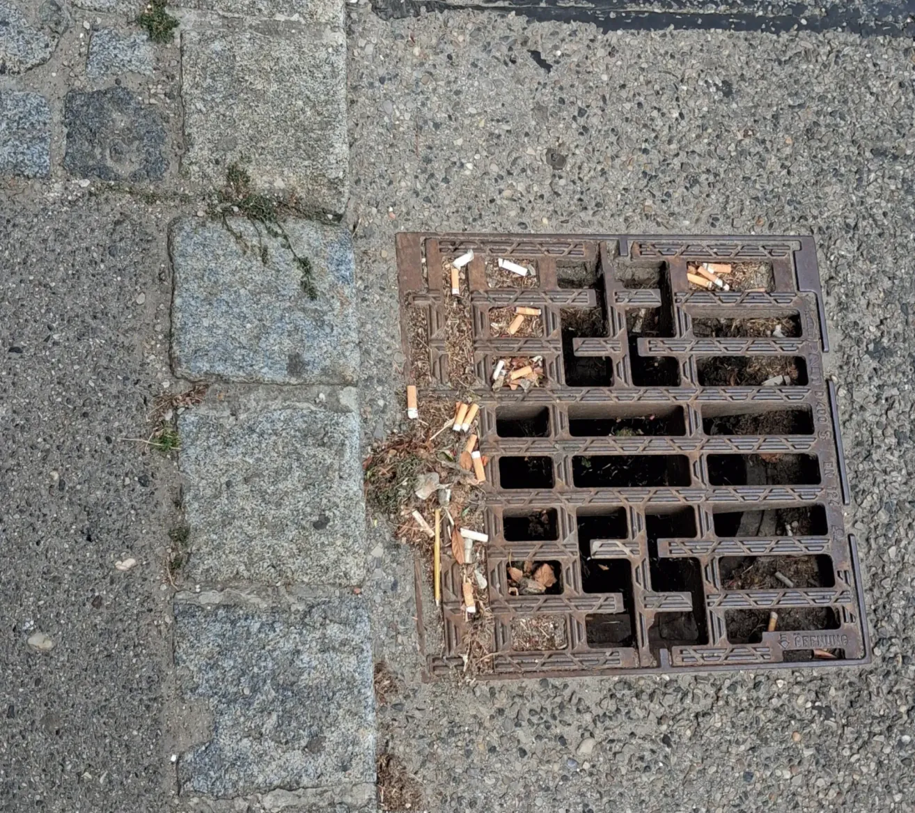 Eine Nahaufnahme eines schmutzigen Kanaldeckels mit Zigarettenkippen und Ablagerungen auf dem Boden.