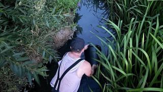Ein Mann in einem weißen Tanktop und schwarzen Trägern beugt sich über einen kleinen Bach und hält einen schwarzen Eimer.