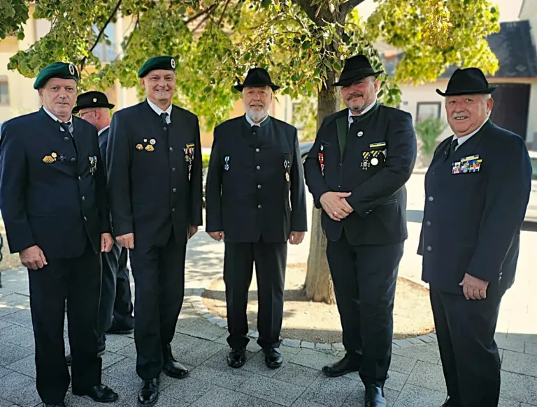Five men in formal attire, including medals, stand under a tree in a park. They are smiling and appear to be posing for a photograph.