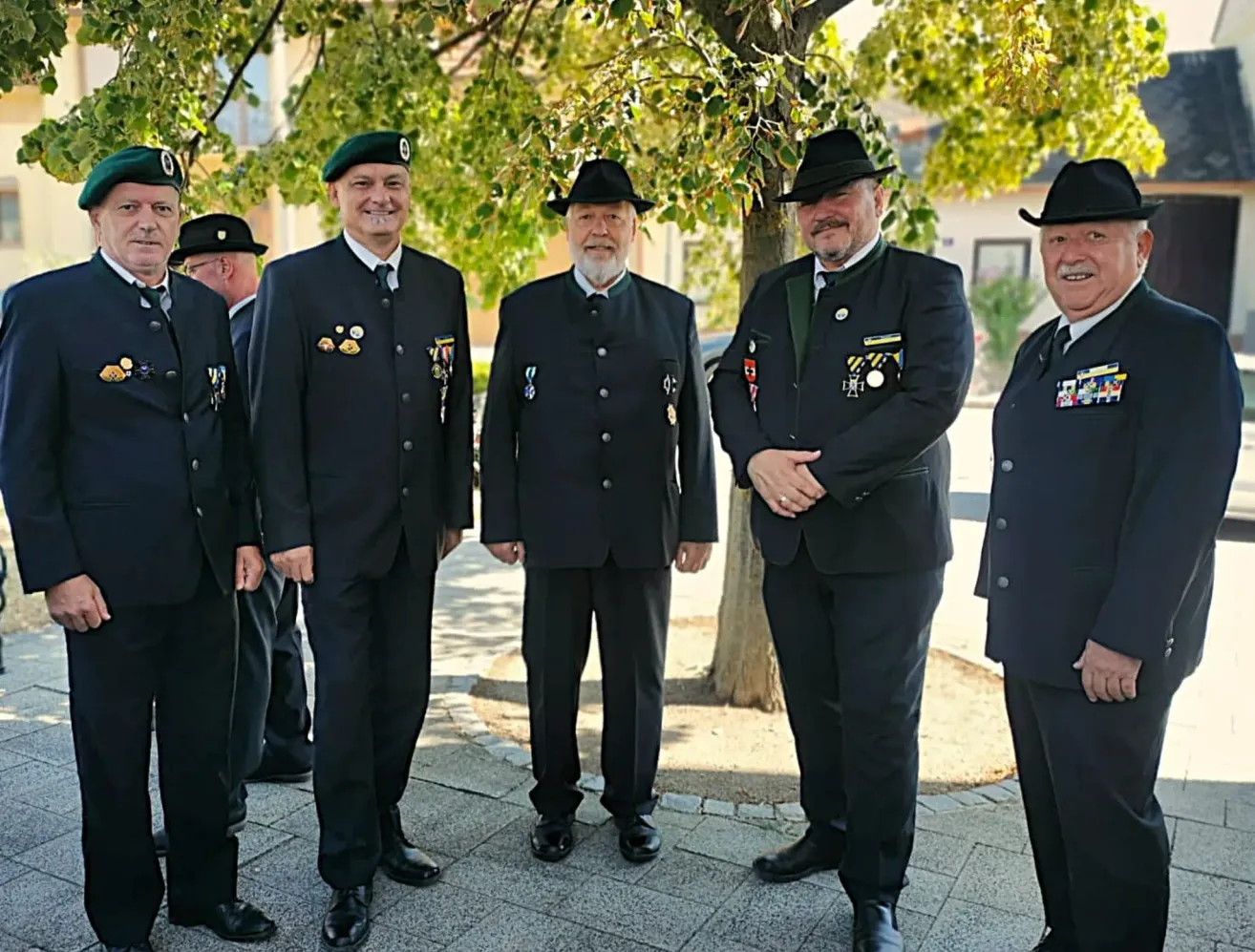 Five men in formal attire, including medals, stand under a tree in a park. They are smiling and appear to be posing for a photograph.