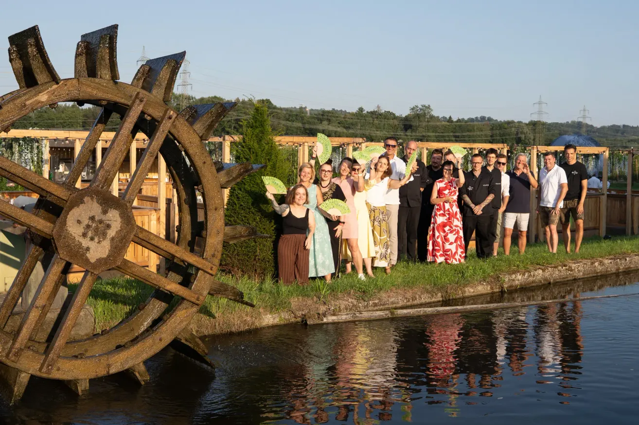 Eine Gruppe von Menschen posiert für ein Foto am Fluss, einige halten Fächer, mit einem großen Wasserrad im Hintergrund.