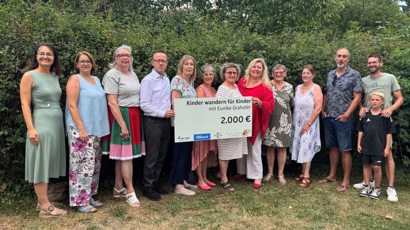 Eine Gruppe Erwachsener steht draußen und hält einen großen Scheck über 2.000 Euro. Der Scheck wird von einer Frau in einer roten Jacke gehalten, und die Gruppe lächelt. Hinter ihnen stehen Büsche und ein klarer blauer Himmel.