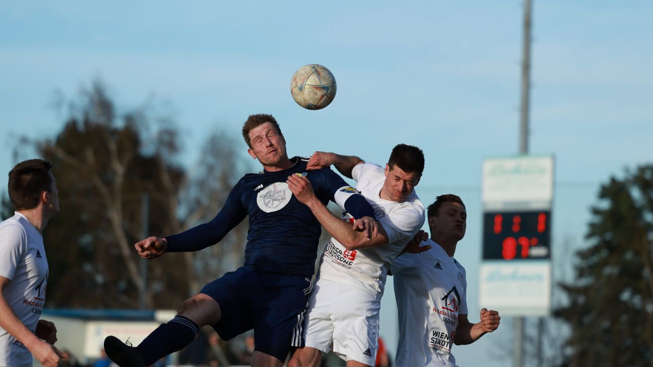 Drei Fußballspieler befinden sich in der Luft, wobei ein Spieler in Blau versucht, den Ball zu köpfen, während zwei Spieler in Weiß versuchen, ihn zu blockieren. Im Hintergrund ist ein klarer blauer Himmel mit einigen Bäumen zu sehen.