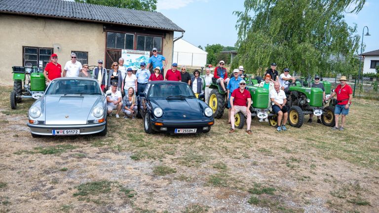 Eine Gruppe von Menschen posiert für ein Foto vor einem Gebäude mit einem Traktor und zwei Autos.