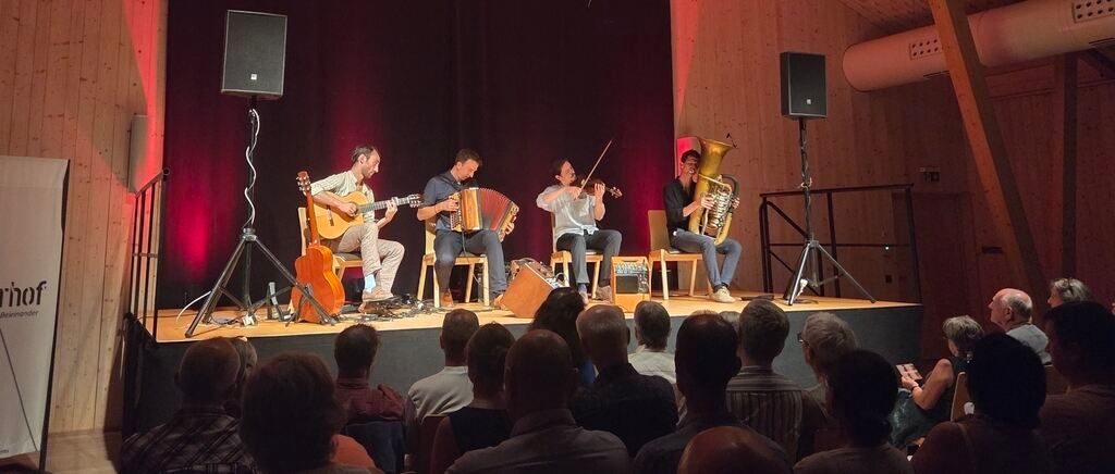 Vier Musiker spielen auf der Bühne mit rotem Vorhang im Hintergrund. Einer spielt Gitarre, ein anderer Geige und zwei weitere Akkordeons. Das Publikum sitzt vor ihnen.