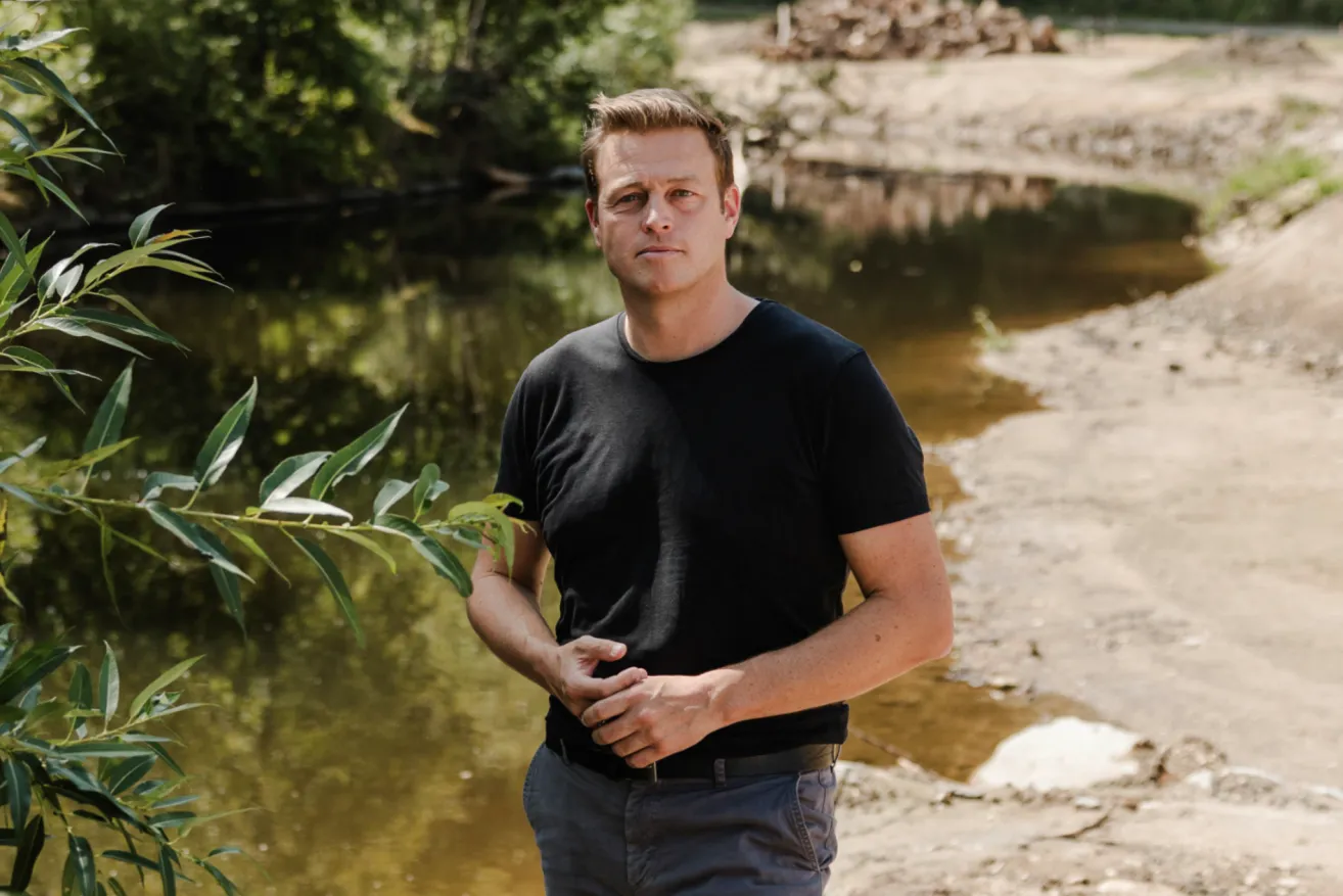 A man stands by a river, wearing a black t-shirt and gray pants, with his hands on his waist, looking to the side.