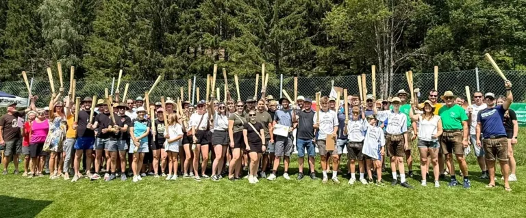 A group of people are standing in a line, holding wooden paddles, and posing for a photo on a grassy field with trees in the background.