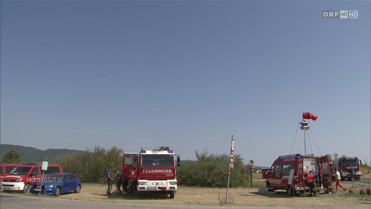 Ein roter Feuerwehrwagen ist am Straßenrand geparkt, neben ihm stehen zwei Personen. Ein blaues Auto ist in der Nähe geparkt und ein Schild steht auf dem Boden. Der Himmel ist klar und blau.