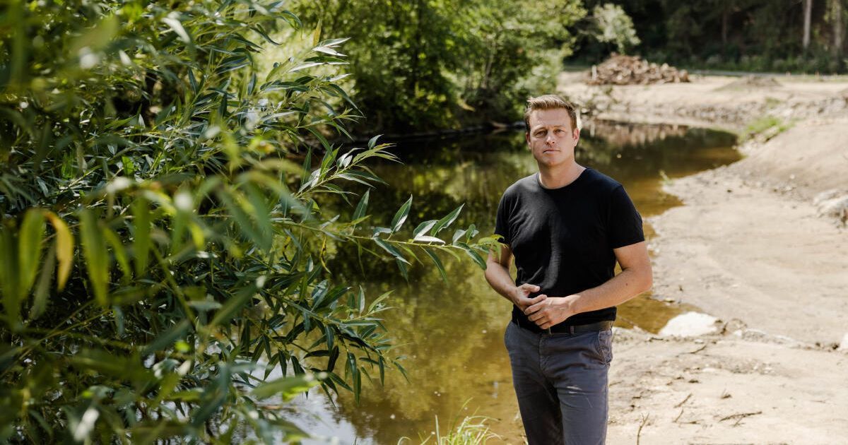 A man stands by a river, wearing a black t-shirt and gray pants, with his hands on his hips.