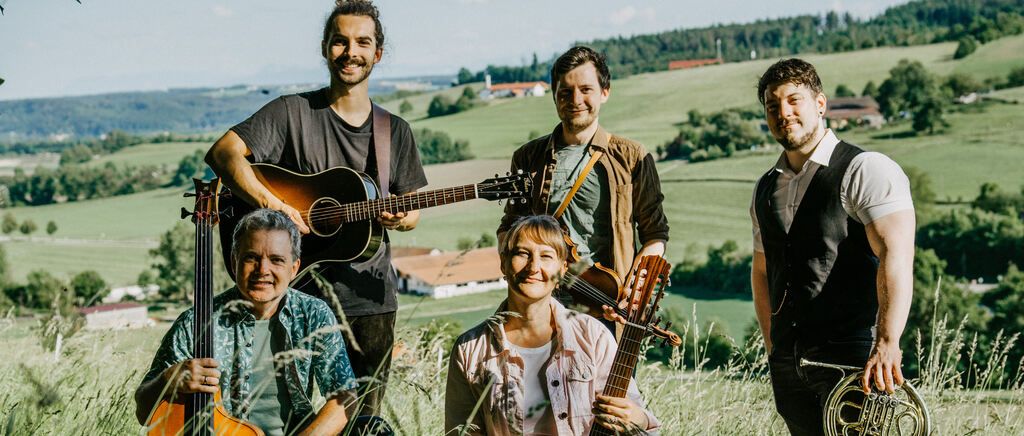 Fünf Musiker, darunter zwei Frauen, stehen in einem Feld und lächeln für ein Foto. Sie halten verschiedene Musikinstrumente, darunter Gitarren, eine Geige und ein Horn. Der Hintergrund zeigt eine grüne Landschaft mit Häusern und Bäumen.