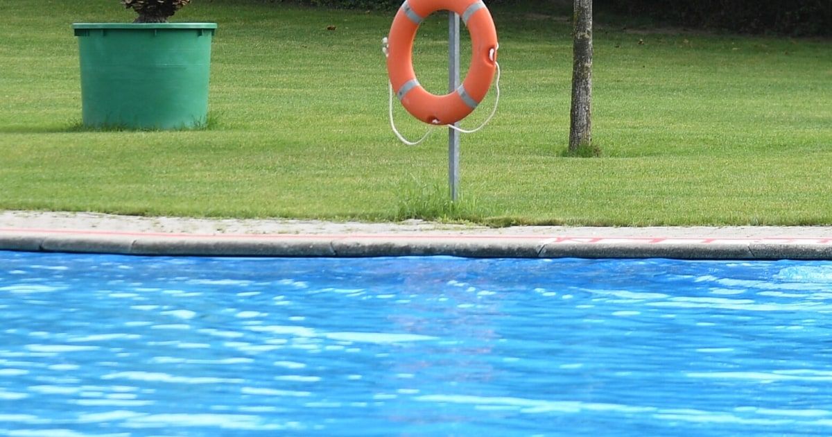 An orange lifebuoy is hanging on a pole near a swimming pool, with a green trash can on the grassy lawn.