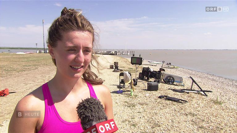 Eine Frau in einem pinkfarbenen Tanktop hält ein Mikrofon mit dem Wort ORF. Sie steht auf einem Strand mit einem Gewässer im Hintergrund. Verschiedene Gegenstände sind verstreut, darunter ein Stuhl, ein Kühler und ein Monitor.