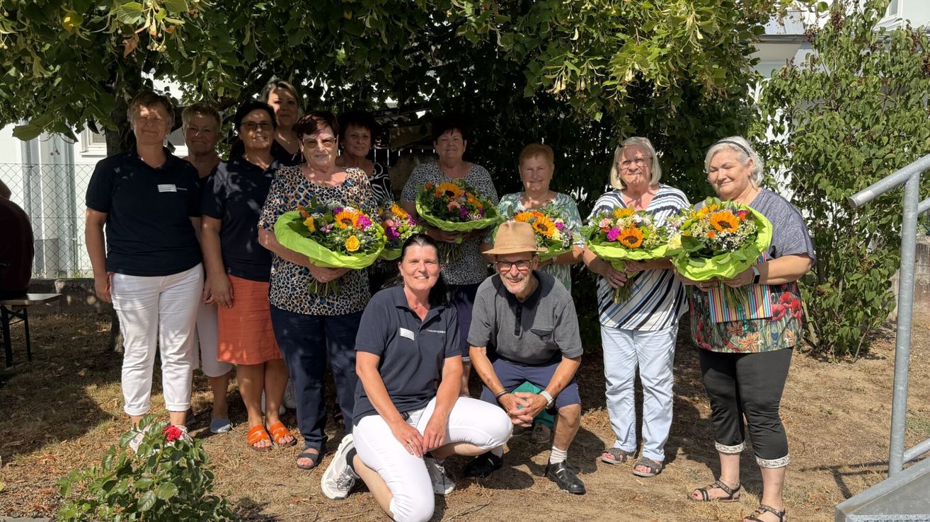 Eine Gruppe älterer Frauen und Männer posiert für ein Foto unter einem Baum, alle halten Blumensträuße. Die Frau in der Mitte kniet, und der Mann neben ihr lächelt.