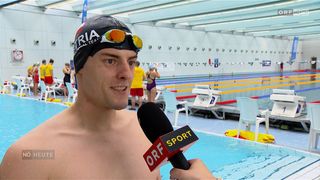 Ein Schwimmer mit einer schwarzen Schwimmmütze, auf der 'Austria' steht, wird von einem Reporter mit einem Mikrofon in einem Hallenbad interviewt.