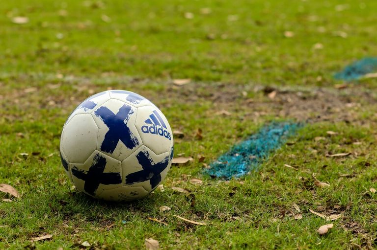 Ein weißer Fußball mit blauer Adidas-Markierung und blauen X-Markierungen auf einem Rasenfeld mit einer blauen Kreidelinie.