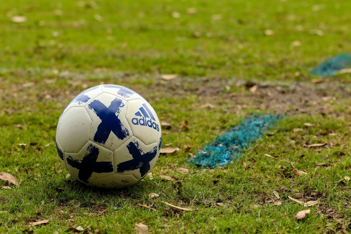 Ein weißer Fußball mit blauer Adidas-Markierung und blauen X-Markierungen auf einem Rasenfeld mit einer blauen Kreidelinie.