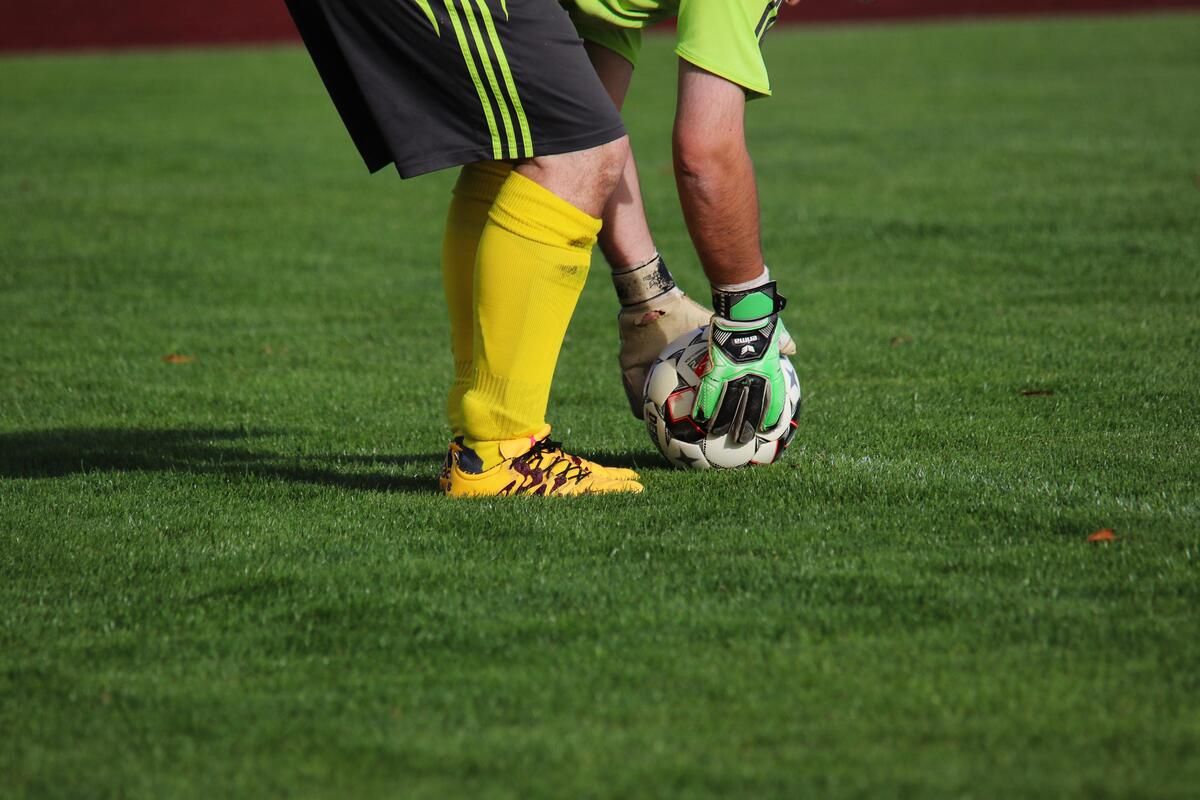 Ein Fußballspieler in gelben Socken und Turnschuhen ist bereit, einen Schuss zu blockieren, während ein Torwart in grünen Handschuhen bereit ist, den Ball zu fangen.
