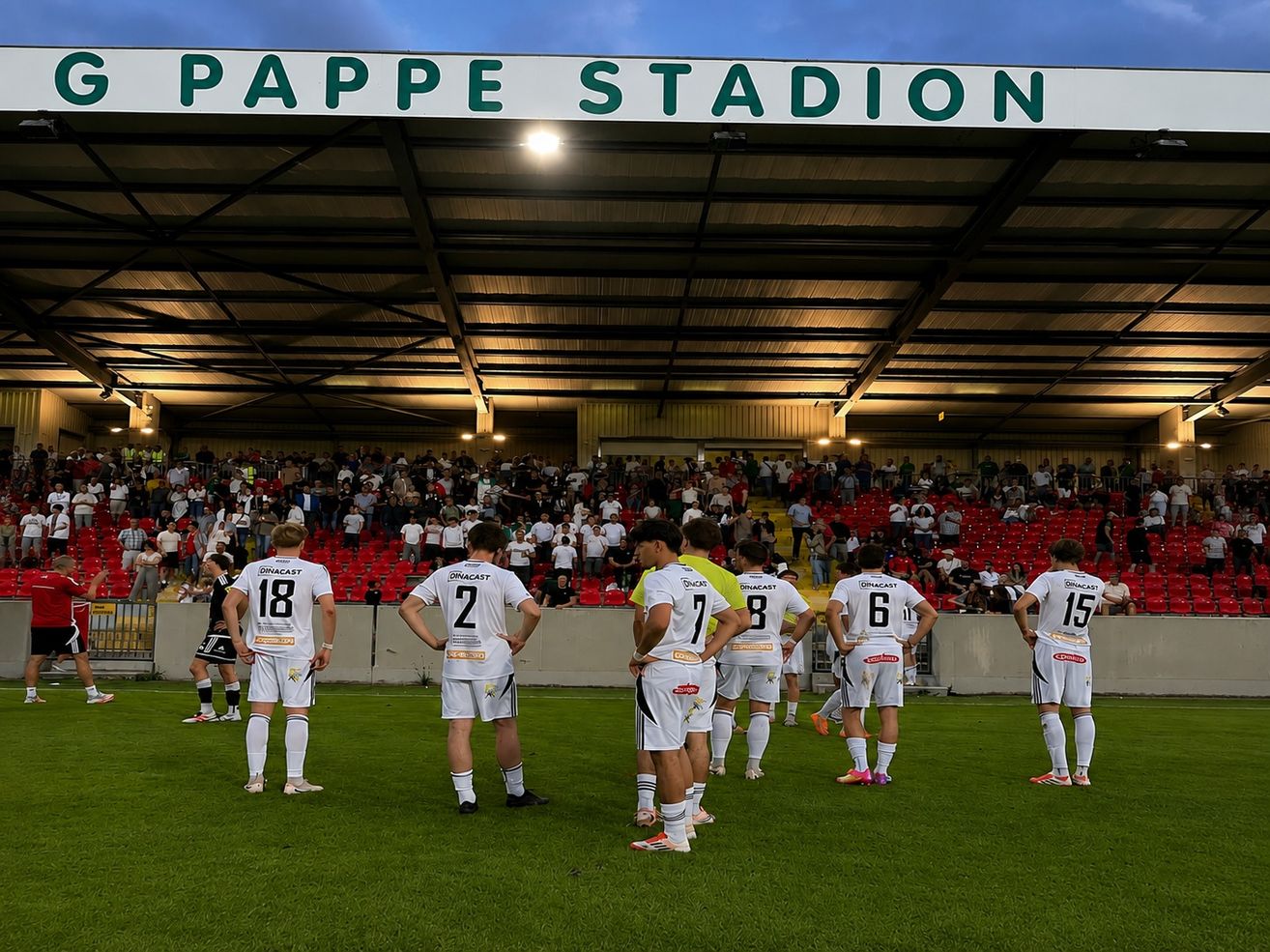Fußballspieler in weißen Trikots stehen auf dem Spielfeld und blicken in Richtung Publikum. Das Stadion heißt Pappe Stadion.