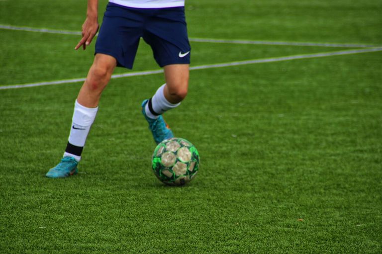 Ein Fußballspieler in blauen Shorts und blauen Schuhen tritt einen grün-braunen Fußball auf einem grünen Feld.