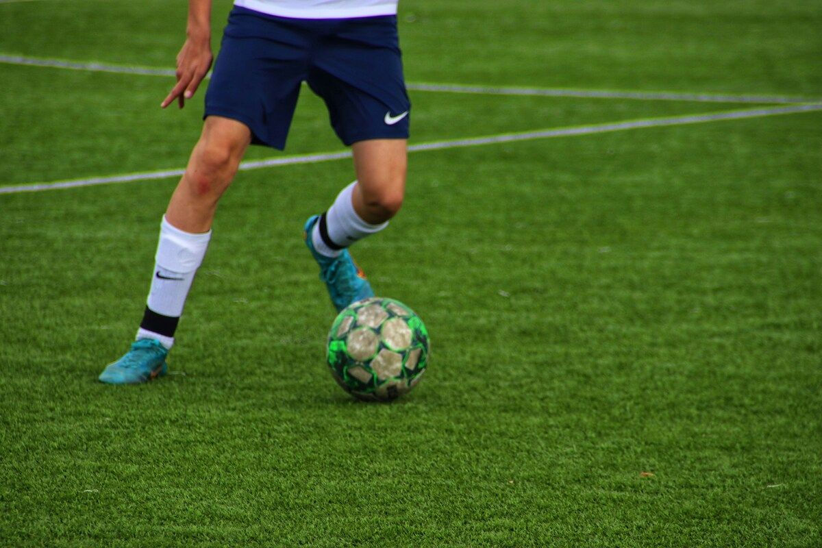 Ein Fußballspieler in blauen Shorts und blauen Schuhen tritt einen grün-braunen Fußball auf einem grünen Feld.