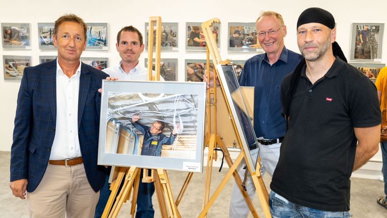 Vier Männer stehen in einem Raum mit gerahmten Bildern an der Wand. Sie halten ein gerahmtes Foto eines Mannes, der an einer Decke arbeitet.