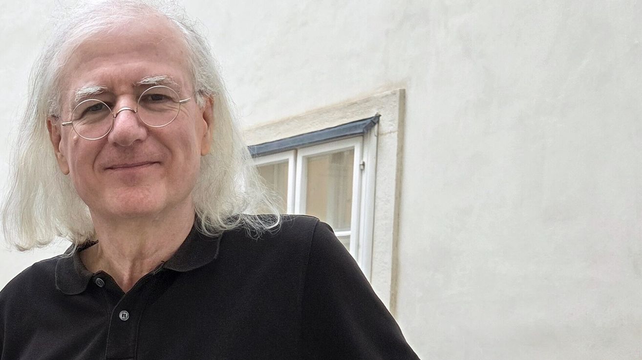 Ein älterer Mann mit langen weißen Haaren und einer Brille steht vor einem weißen Gebäude. Er trägt ein schwarzes Hemd und lächelt.