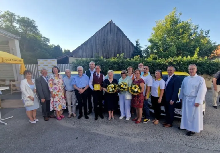 Eine Gruppe von Menschen in formeller Kleidung, einige mit Blumensträußen, steht vor einem gelben Fahrzeug mit einem Scheunengebäude im Hintergrund.