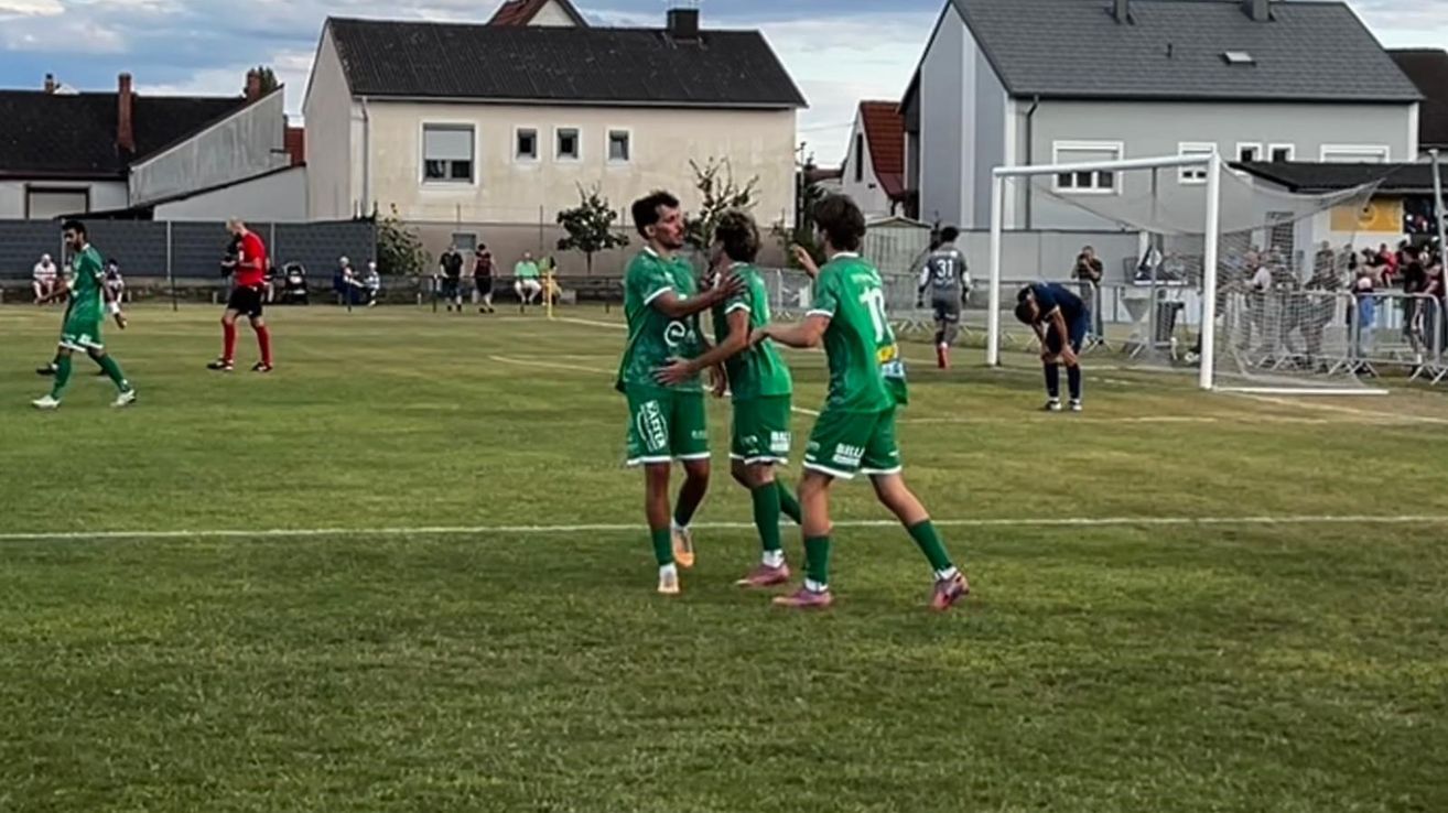 Drei Fußballspieler in grünen Trikots feiern ein Tor auf einem Spielfeld mit einem Torpfosten im Hintergrund.