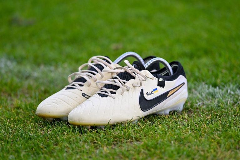 Ein Paar weißer Fußballschuhe mit schwarzen und goldenen Akzenten und dem Wort 'Maxi' an der Seite. Die Schuhe liegen auf einem grünen Rasen.