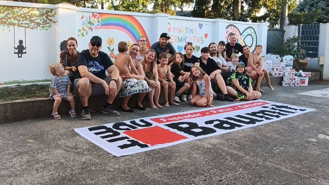 Eine Gruppe von Menschen, einschließlich Erwachsener und Kindern, sitzen und stehen auf einer Betonfläche und posieren für ein Foto. Ein großer Banner mit dem Wort 'Bau' liegt auf dem Boden. Dahinter befindet sich eine Wand mit einem Regenbogen und anderen Zeichnungen.