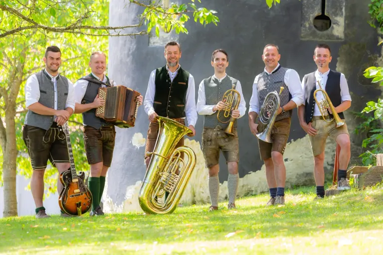 Fünf Männer in traditioneller bayerischer Kleidung, jeder mit einem anderen Musikinstrument, stehen auf einem Rasen. Sie lächeln und scheinen für ein Foto zu posieren.
