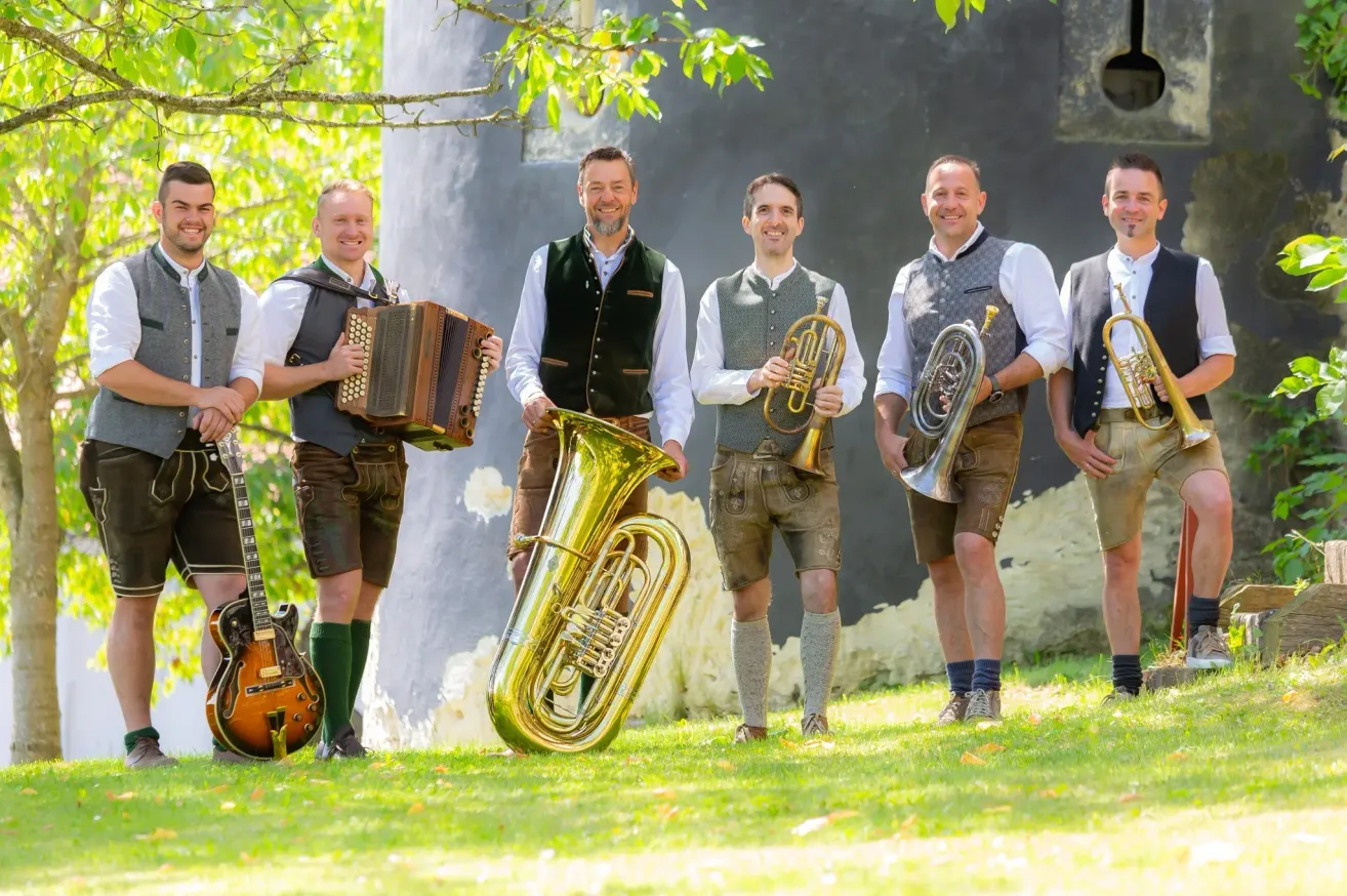 Fünf Männer in traditioneller bayerischer Kleidung, jeder mit einem anderen Musikinstrument, stehen auf einem Rasen. Sie lächeln und scheinen für ein Foto zu posieren.